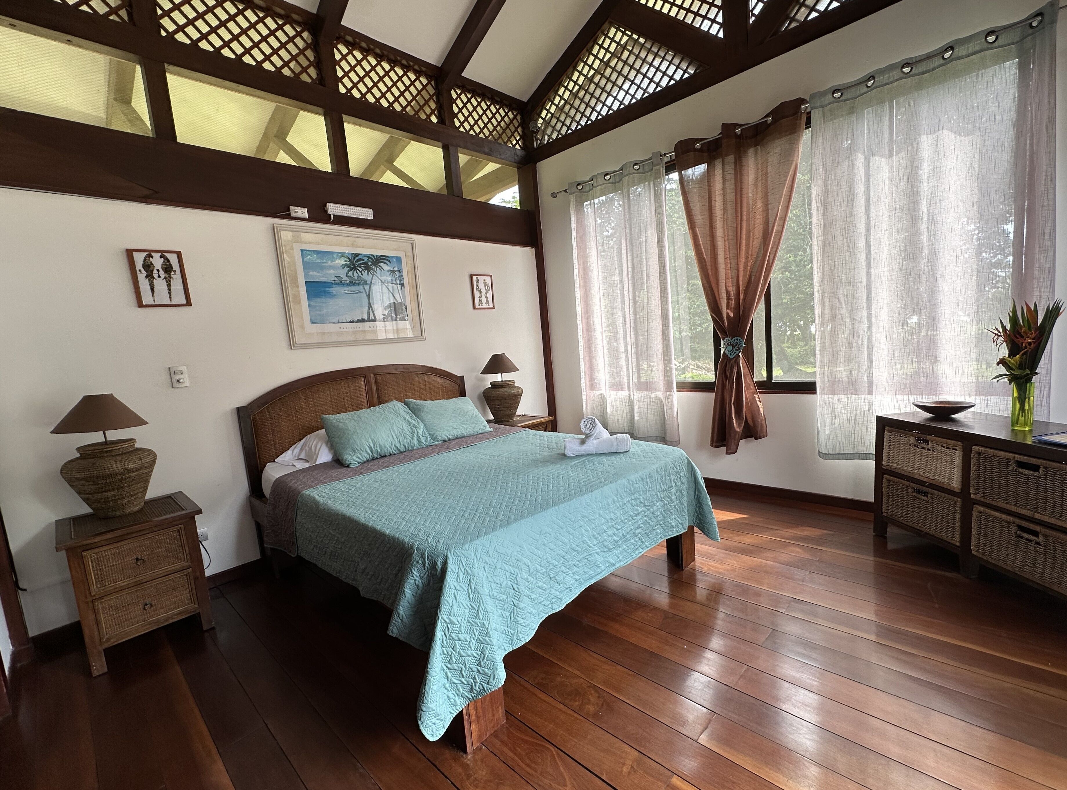 A bedroom with a bed, wooden floors, a nightstand with a lamp, a dresser, and a window with curtains.