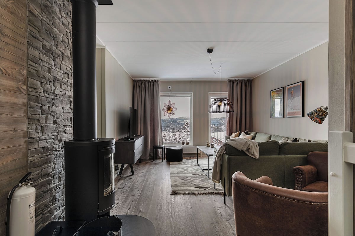 Ein gemütliches Wohnzimmer mit einer Steinwand, einem schwarzen Ofen, einer Couch und Blick durch ein Fenster.