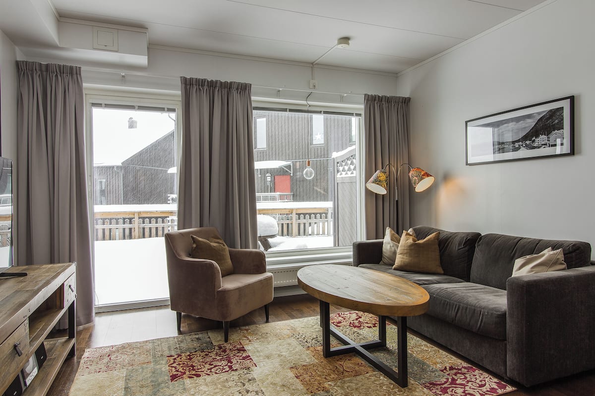 Ein modernes Wohnzimmer mit Sofa, Sessel und Couchtisch, großen Fenstern mit Vorhängen und Blick auf ein Gebäude.
