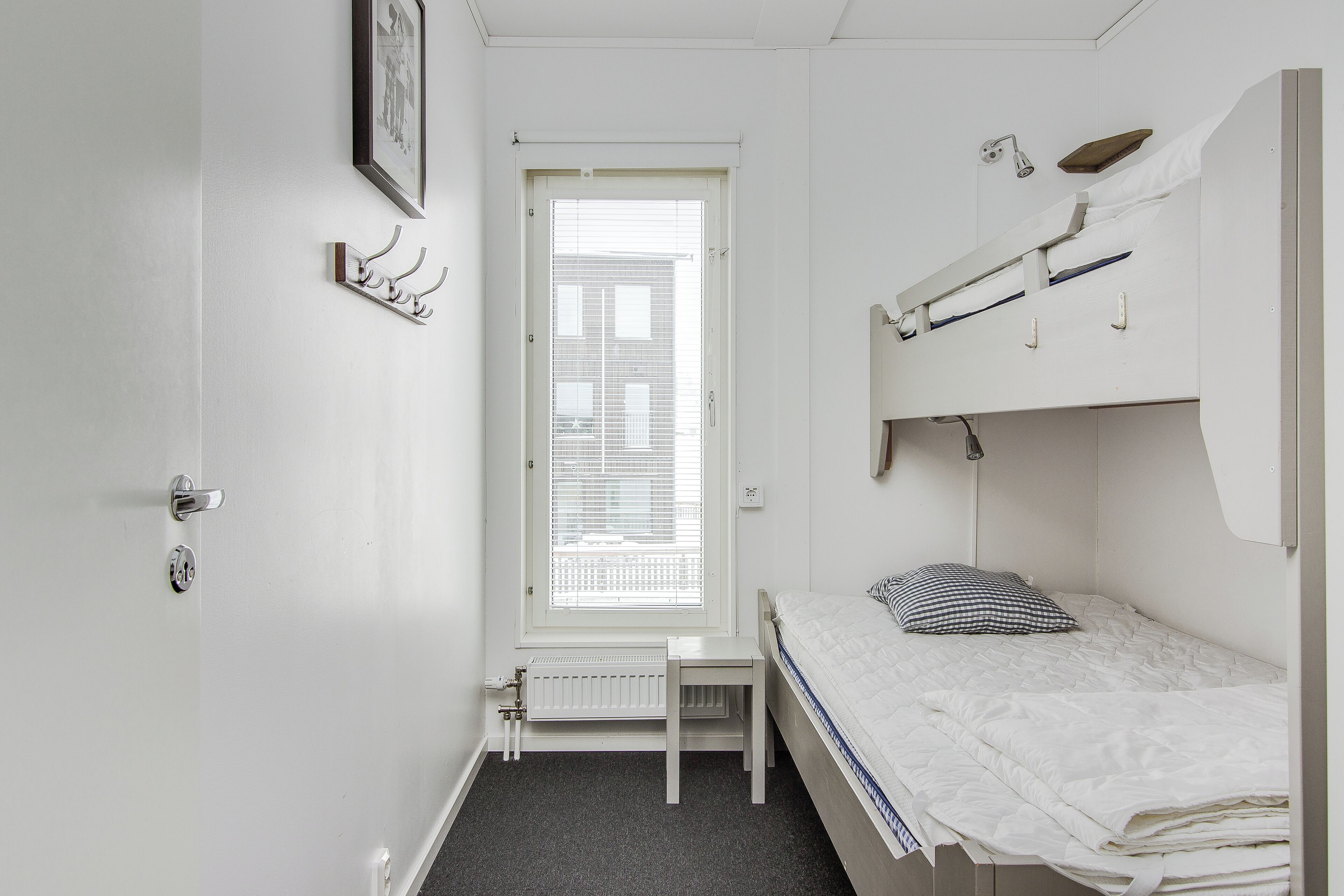 A small, white room with a single bed, a radiator, a window with blinds, a framed picture on the wall, and a door with a handle.