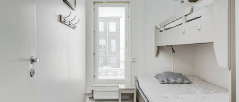 Una stanza piccola e bianca con un letto singolo, un termosifone, una finestra con persiane, un quadro appeso al muro e una porta con maniglia.