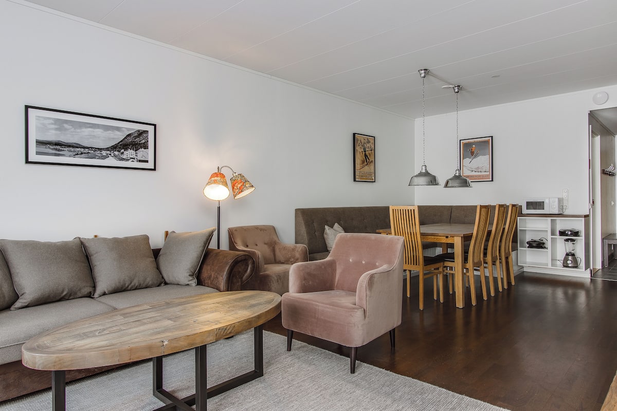 Ein modernes Wohnzimmer mit Sofa, Sessel, Esstisch und Stühlen. An den Wänden hängen gerahmte Bilder und auf dem Boden steht eine Stehlampe.