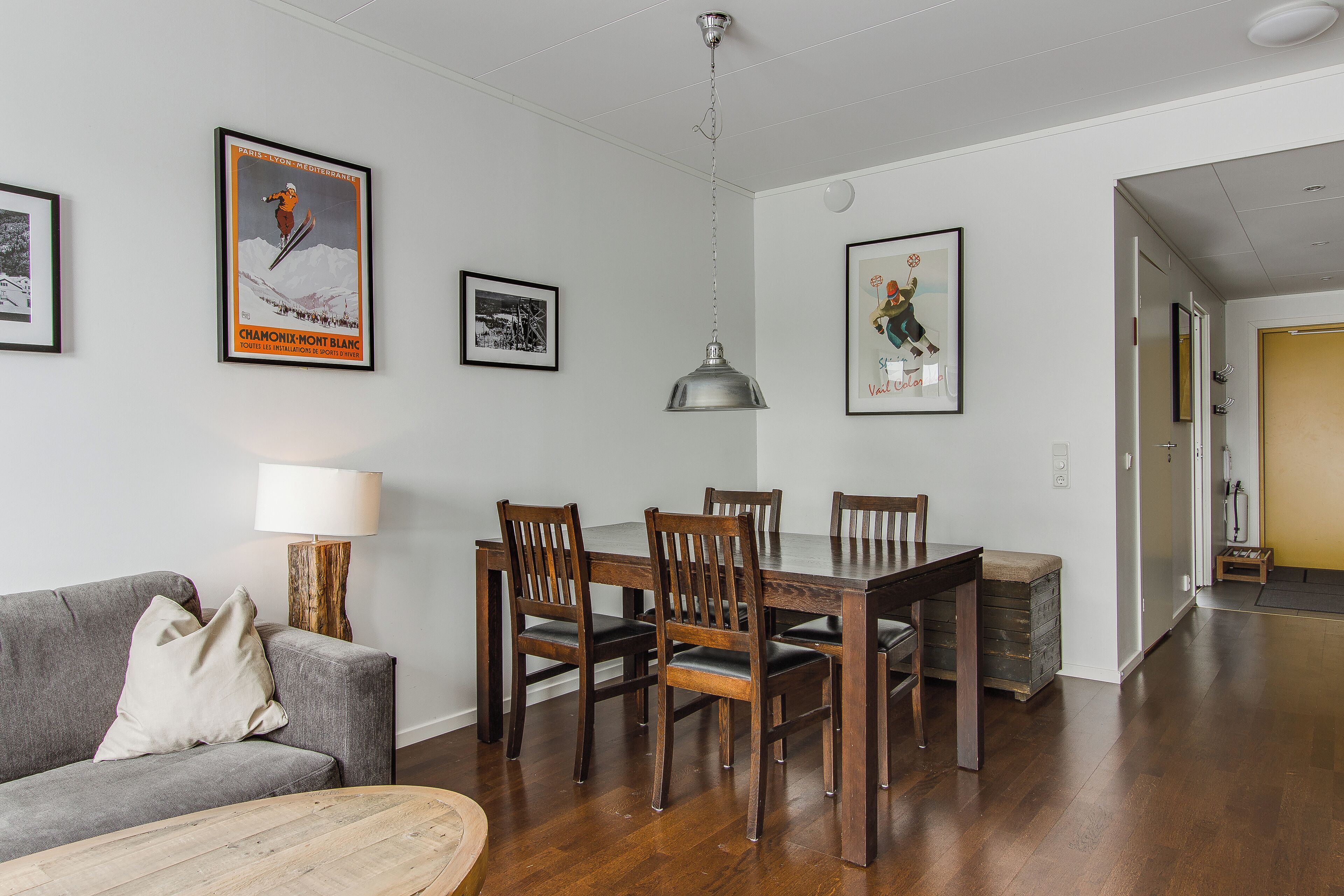 Sala de estar moderna com sofá cinzento, mesa de jantar de madeira e cadeiras. Há quadros nas paredes, um lustre pendurado e chão de madeira.