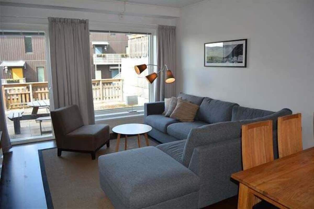 Ein modernes Wohnzimmer mit einem grauen Sofa, einem Holztisch und einem Sessel. Ein großes Fenster mit Vorhängen, eine Hängelampe und ein gerahmtes Bild sind ebenfalls zu sehen.