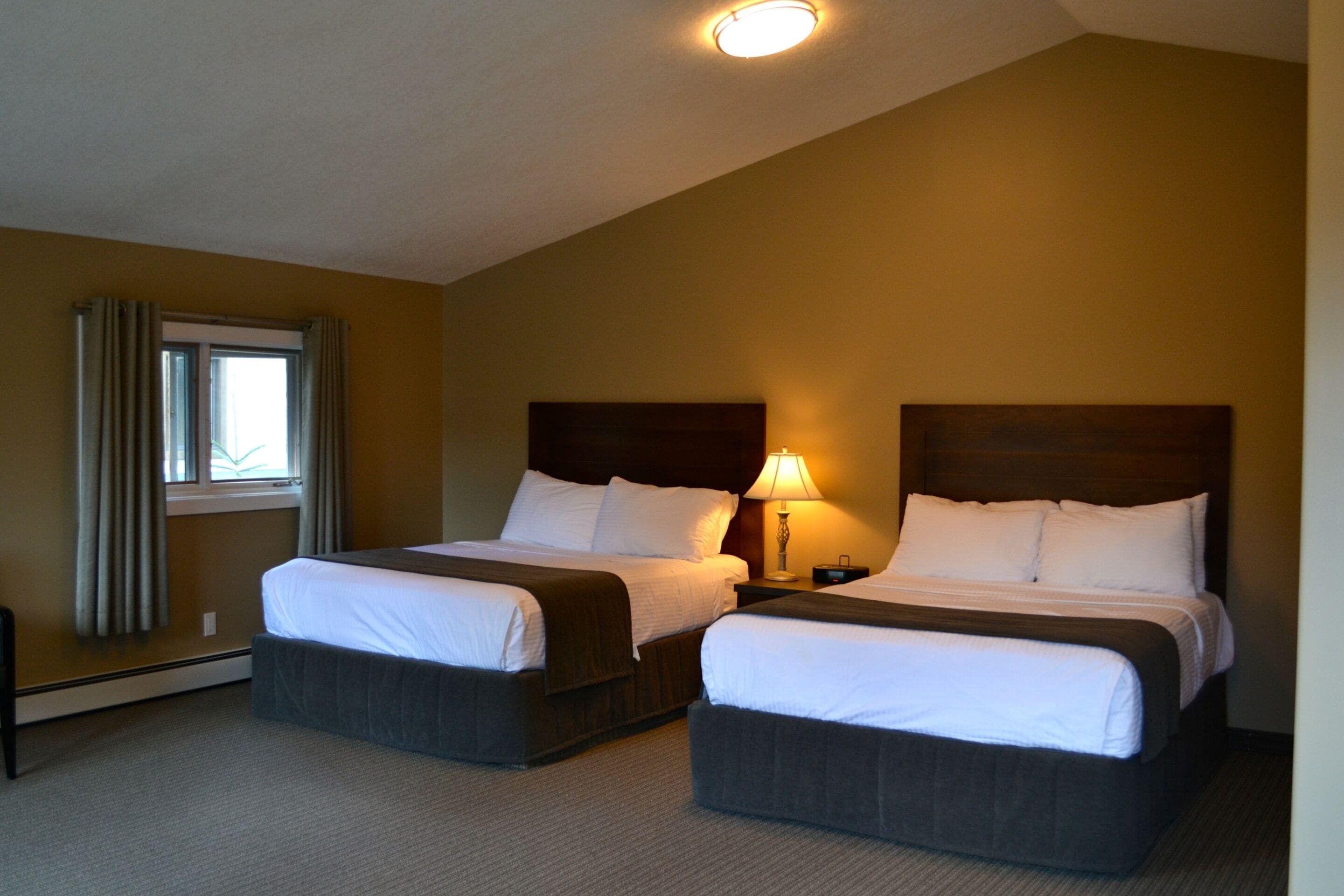 Une chambre d’hôtel avec deux lits, une fenêtre avec des rideaux, une table de chevet avec une lampe et un luminaire au plafond.