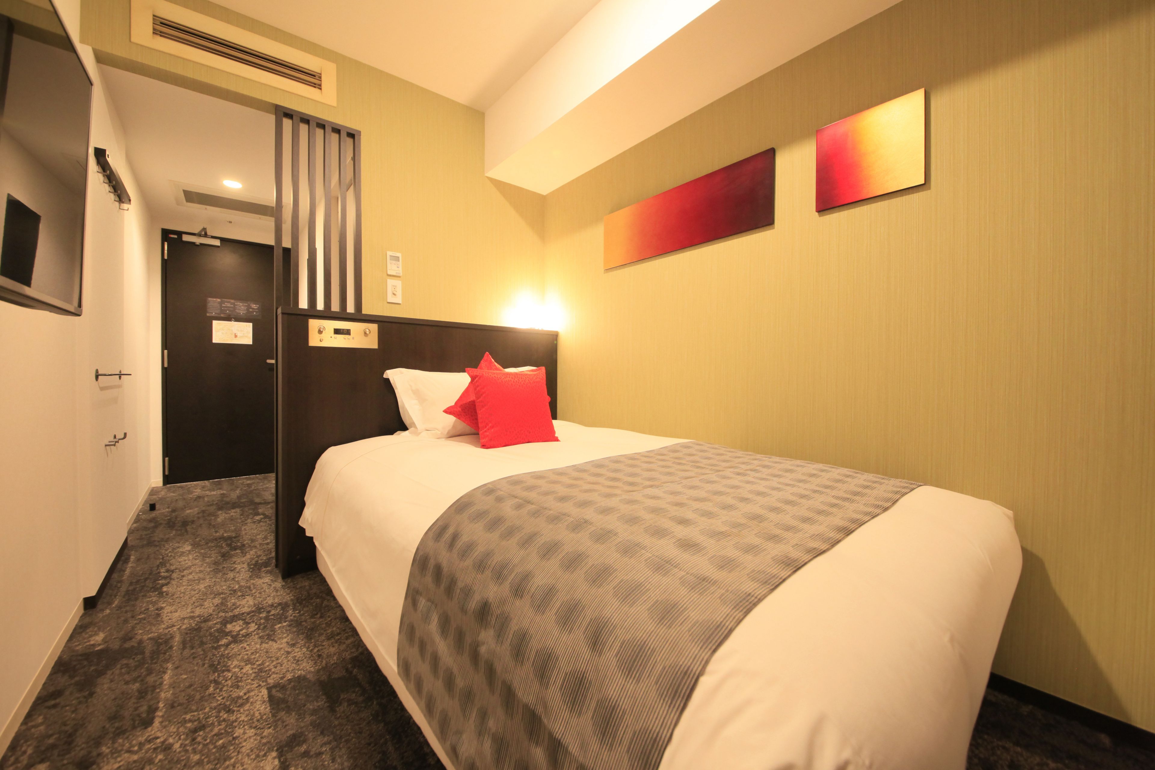 A hotel room with a bed, a door, a wall with three red panels, and a carpeted floor.