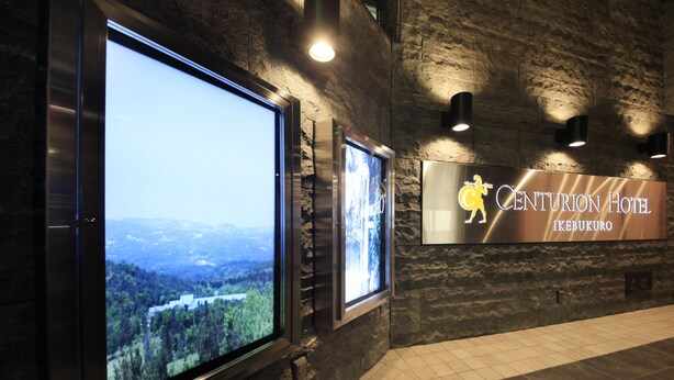 A wall with two large digital screens displaying scenic views, a sign for Centurion Hotel in Ikebukuro, and a tiled floor. - Centurion Hotel Ikebukuro (Tokyo)