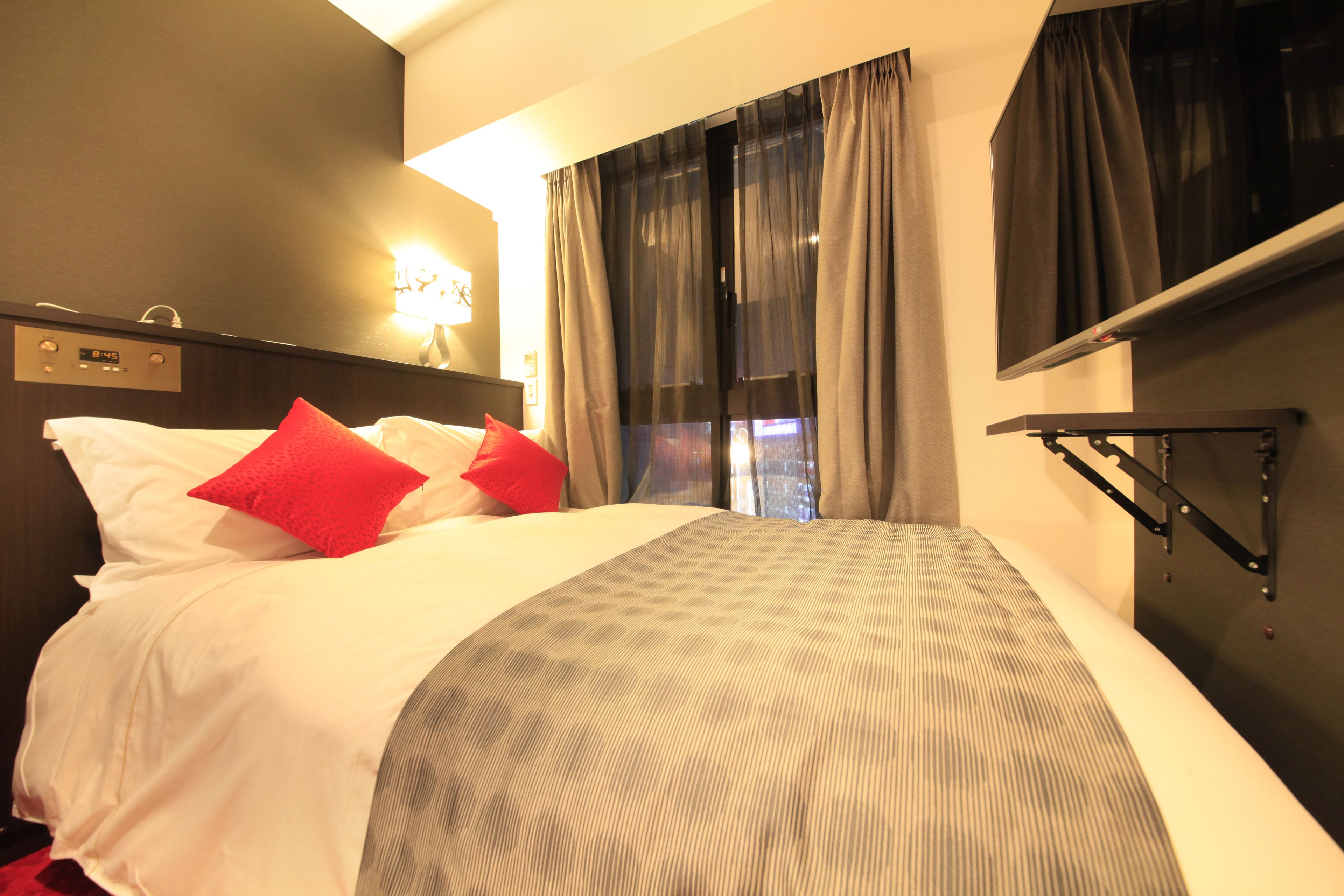 A hotel room with a bed, a nightstand, a wall clock, and a window with curtains.