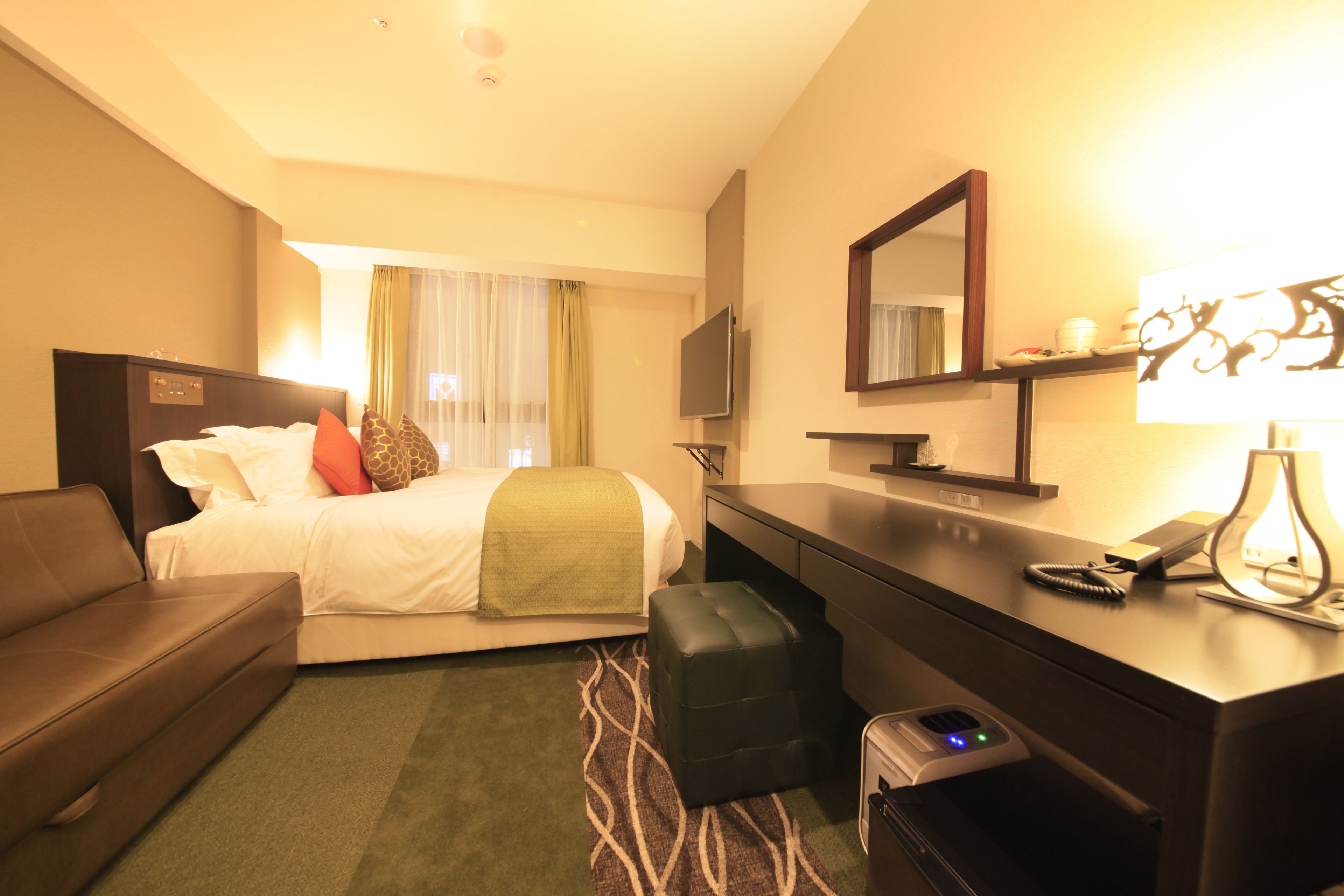 A hotel room with a bed, a desk, a chair, a sofa, a TV, a mirror, and a lamp.