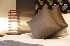A neatly made bed with white linens and two brown pillows, a tall, modern bedside lamp, and a patterned wall in the background. - Centurion Hotel Ikebukuro (Tokyo)