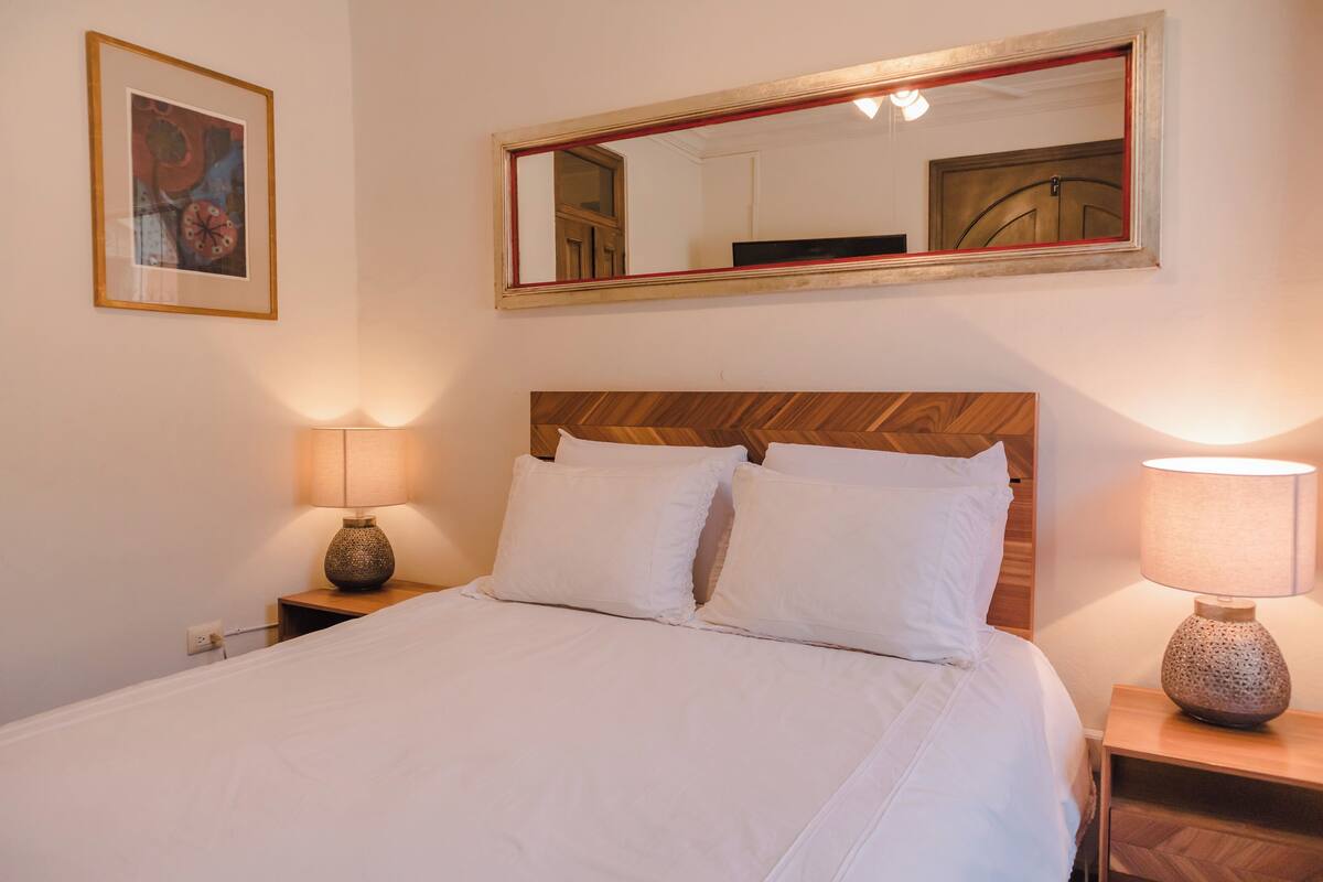 Un lit bien fait, avec du linge de lit blanc, une tête de lit en bois, deux lampes de chevet et un miroir encadré au-dessus du lit.