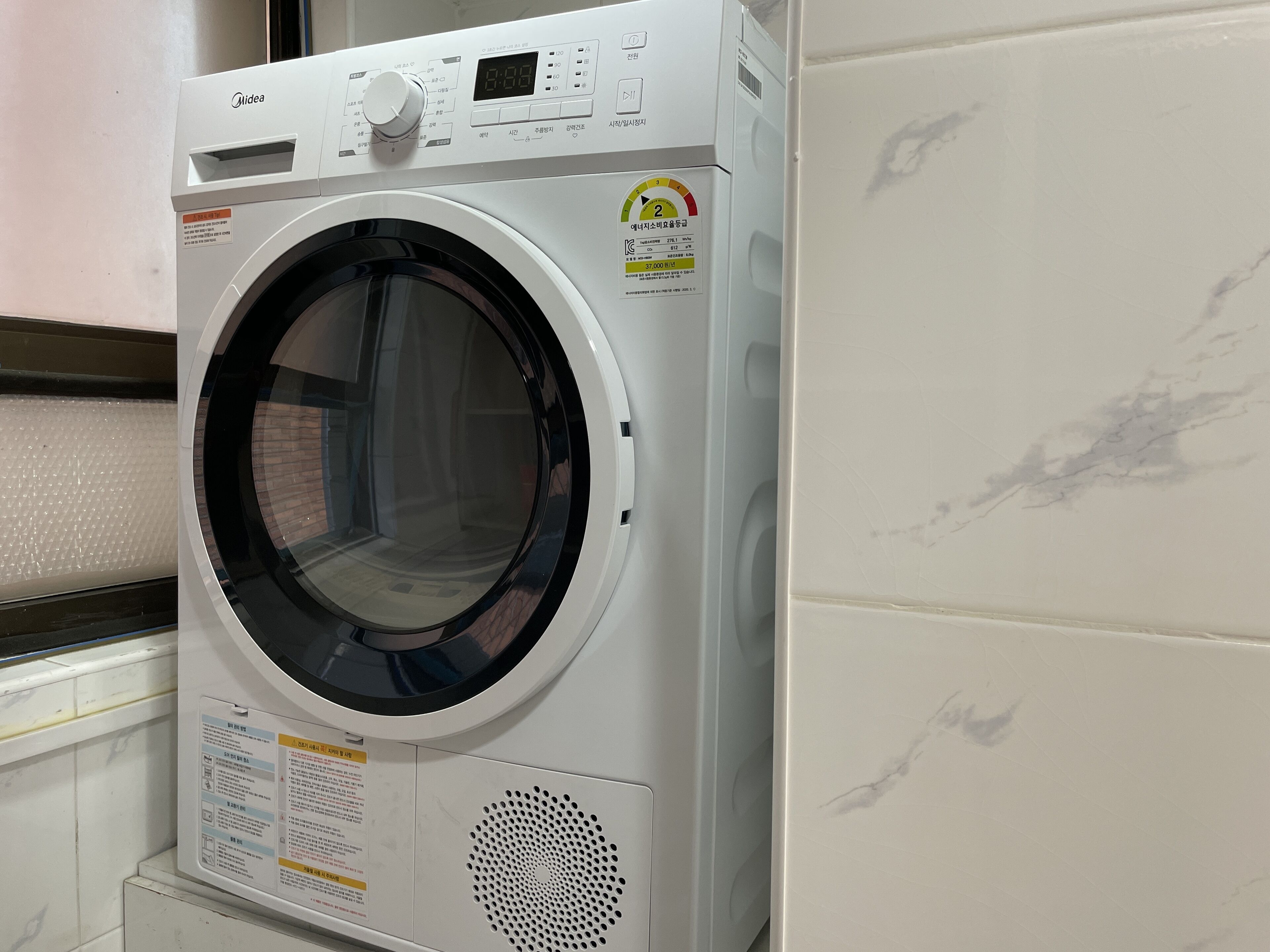 A front-loading washing machine with a digital display, control panel, and a safety label.