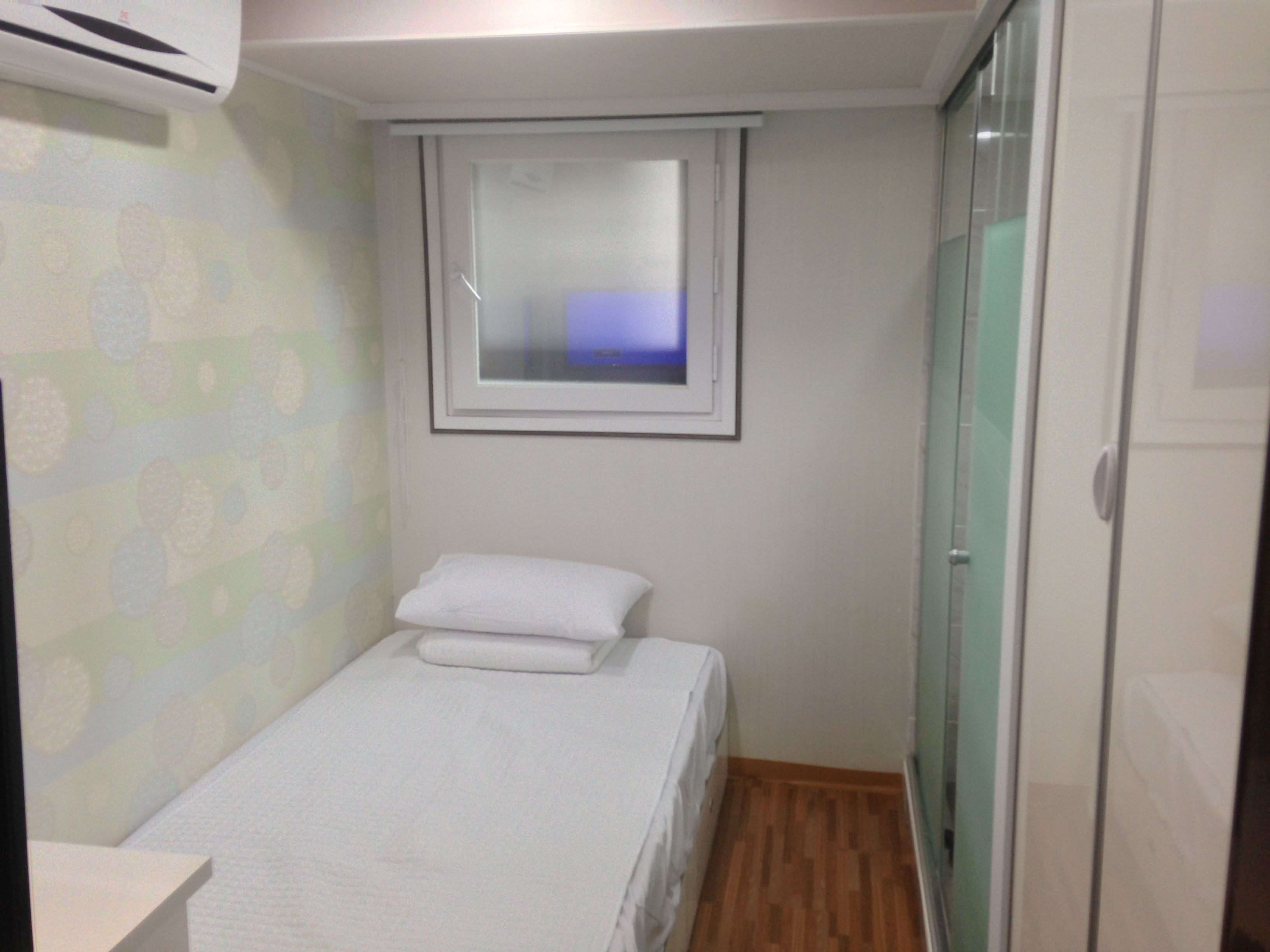 A small, single-bed room with a window, a wooden floor, and a patterned wall.