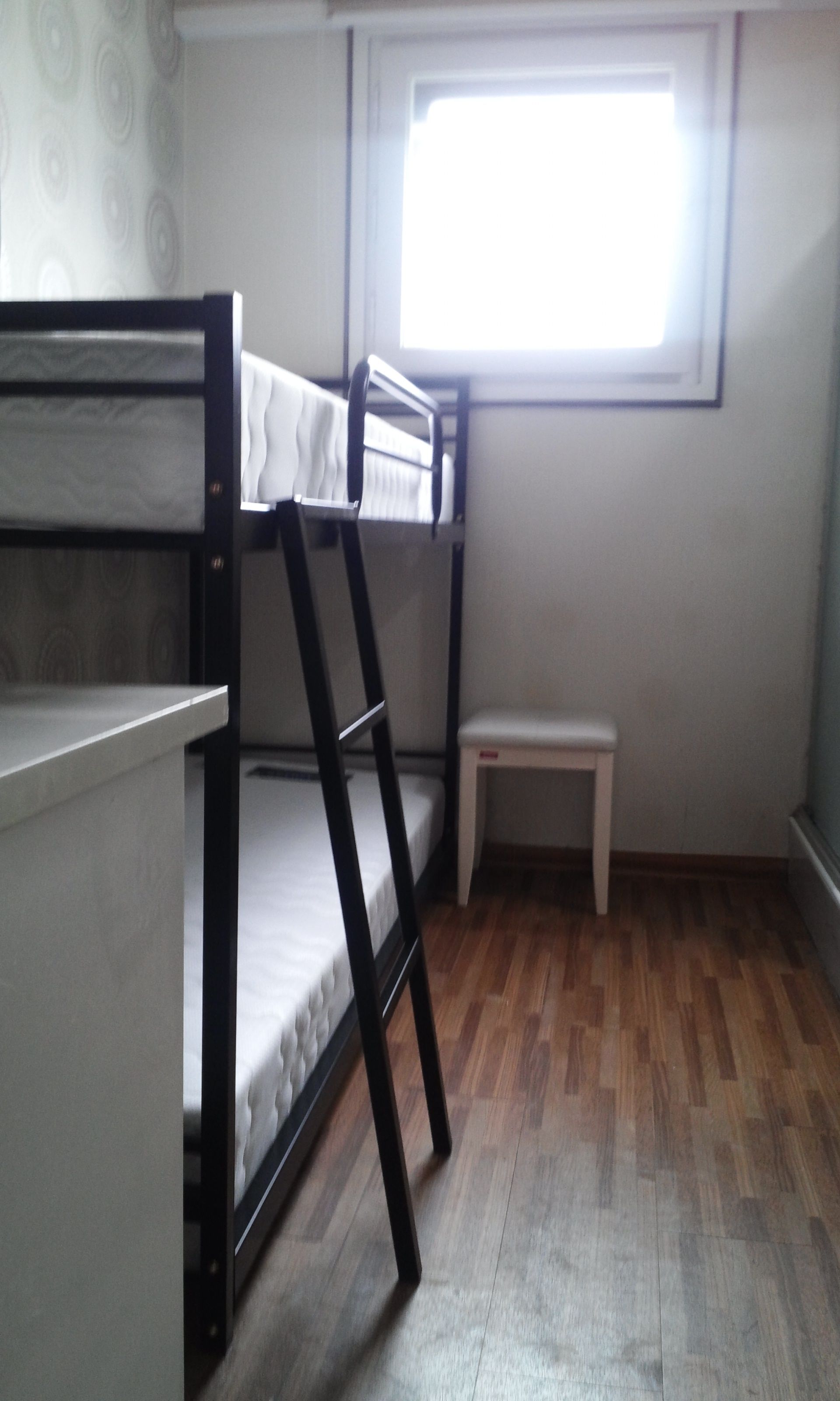 Quarto com beliche, piso de madeira, um pequeno banco branco e janela com persianas.