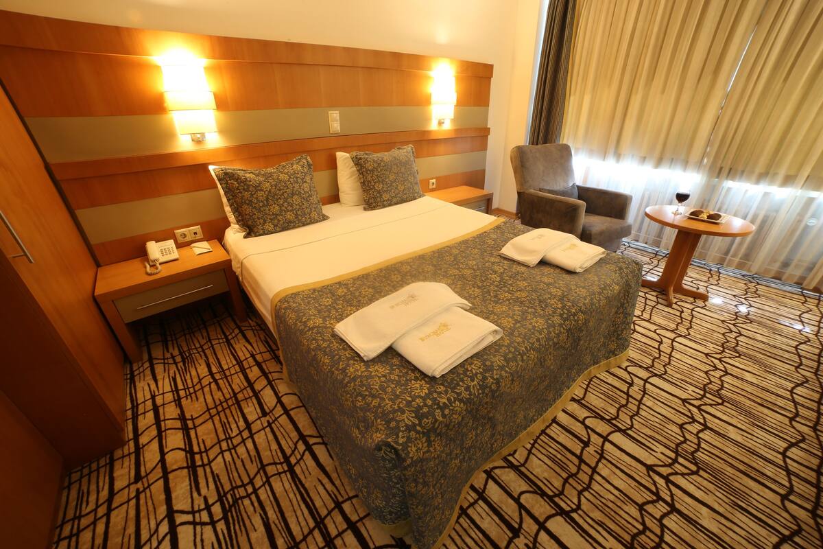 Habitación de hotel con una cama grande, dos mesitas de noche, una silla, una pequeña mesa con una copa de vino y una alfombra estampada.
