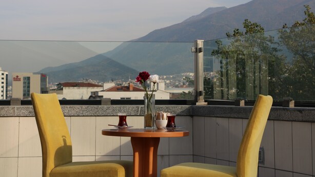 A balcony with two yellow chairs, a round table with a vase of flowers, and a cityscape view in the background. - Burçman Hotel (Bursa)