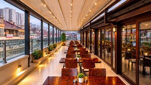A long, narrow indoor dining area with wooden tables and chairs, large windows, and a city view. - Burçman Hotel (Bursa)