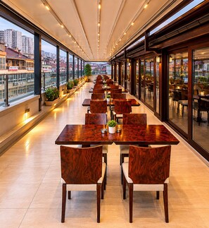 A long, narrow indoor dining area with wooden tables and chairs, large windows, and a city view. - Burçman Hotel (Bursa)