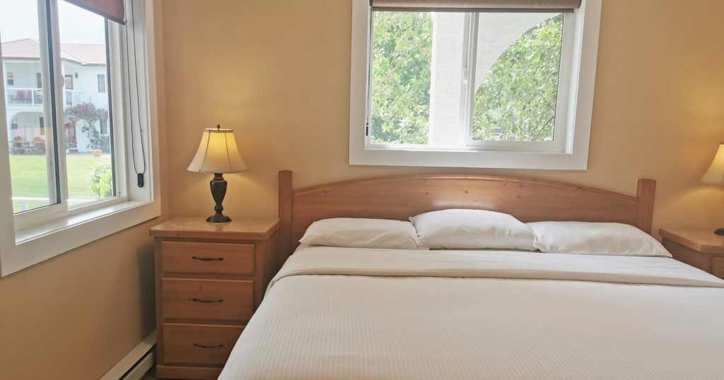 Ein Schlafzimmer mit hölzernem Kopfteil, einem Bett mit weißer Bettwäsche, einem Nachttisch mit Lampe und einem Fenster mit Blick auf Grünflächen.