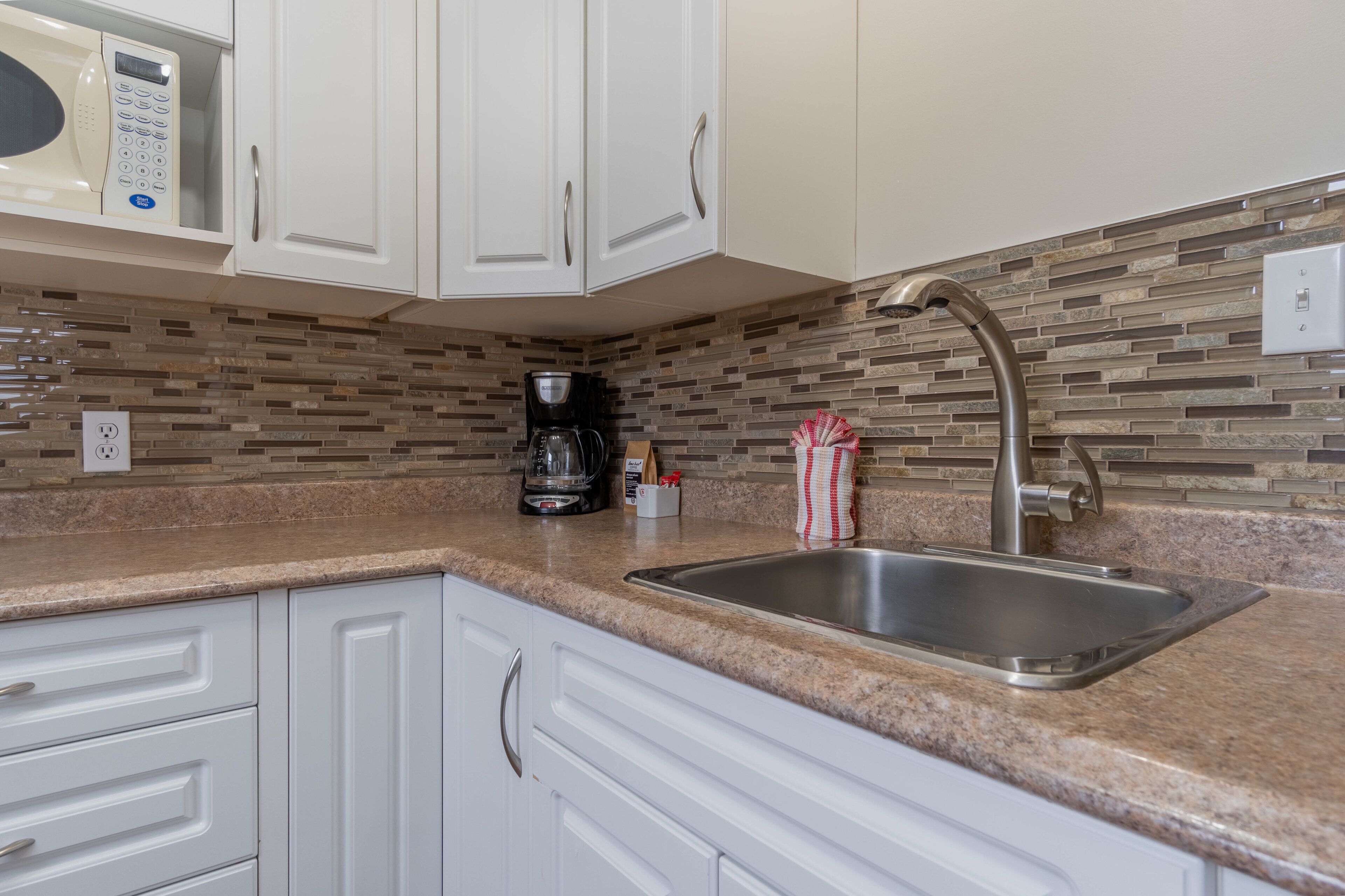 Una cocina con gabinetes blancos, un lavamanos de acero inoxidable, una cafetera y un microondas.