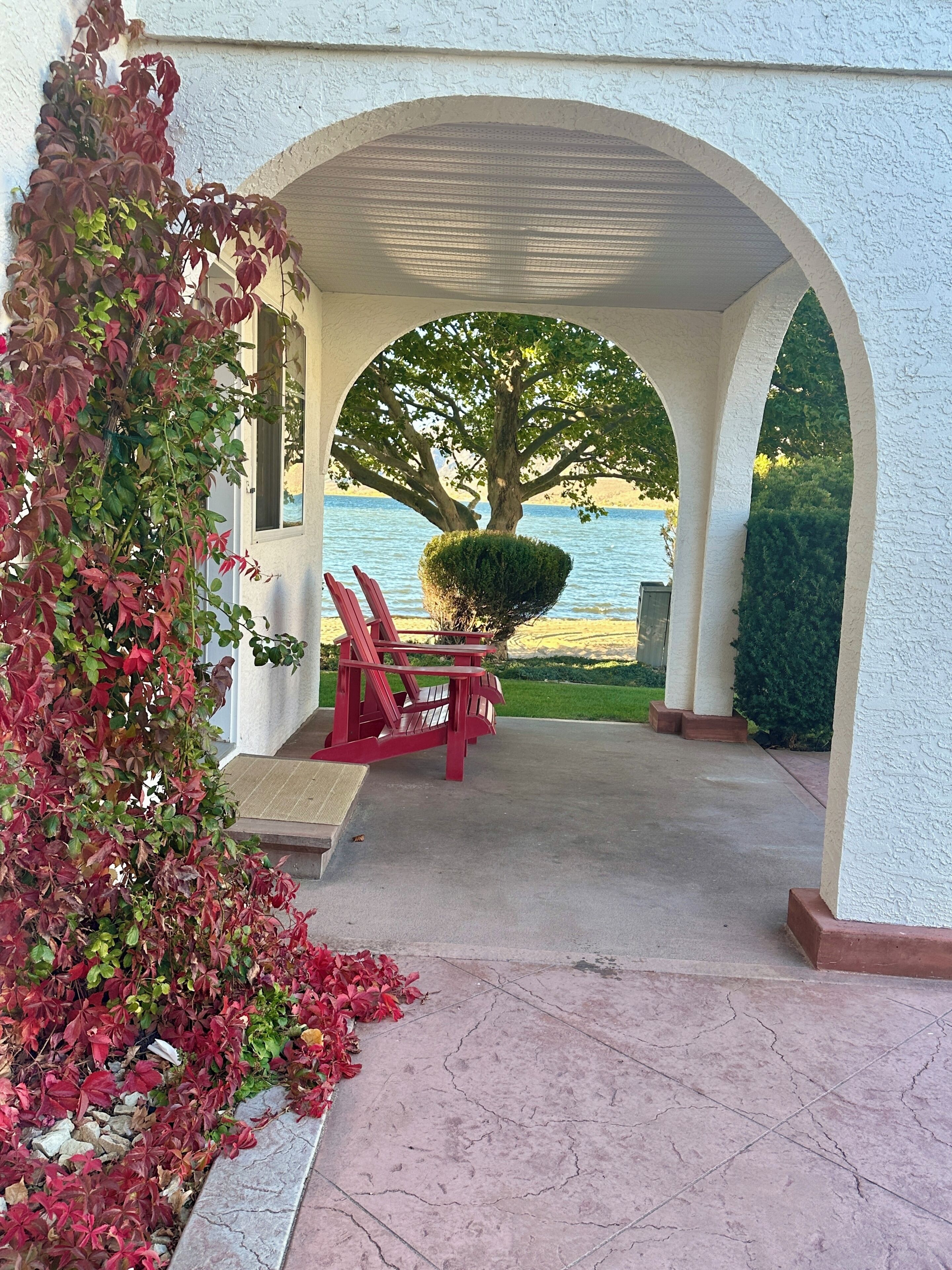 A covered outdoor seating area with a view of a lake, a tree, and a red bench.