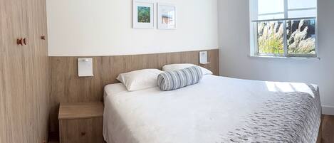 Ein Schlafzimmer mit einem Bett, einem Nachttisch, einem Fenster mit Blick auf Pflanzen und zwei gerahmten Bildern an der Wand.