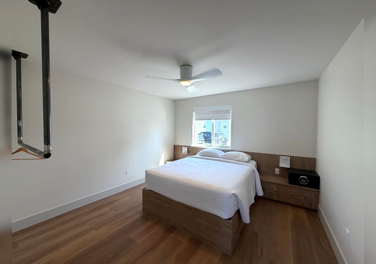 Un dormitorio con una cama, un ventilador de techo, una ventana y una mesita de noche.