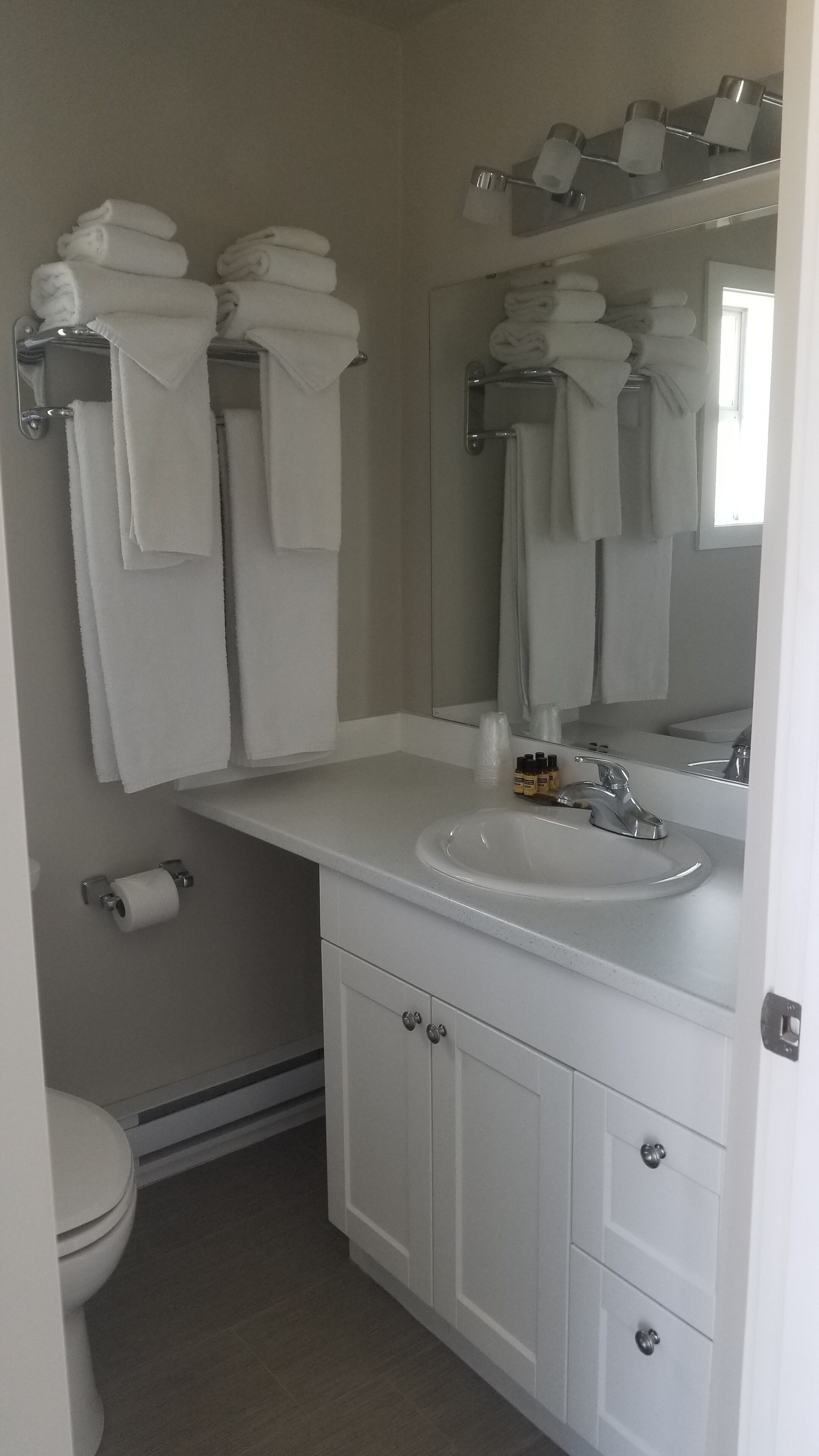 Une salle de bain avec un lavabo blanc, un miroir et des porte-serviettes. On y trouve des articles de toilette sur le comptoir et un rouleau de papier toilette accroché au mur.