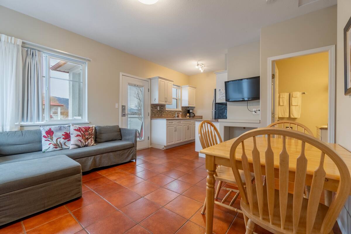 A modern living room with a sofa, a dining table with chairs, and a kitchen area with cabinets and a TV.