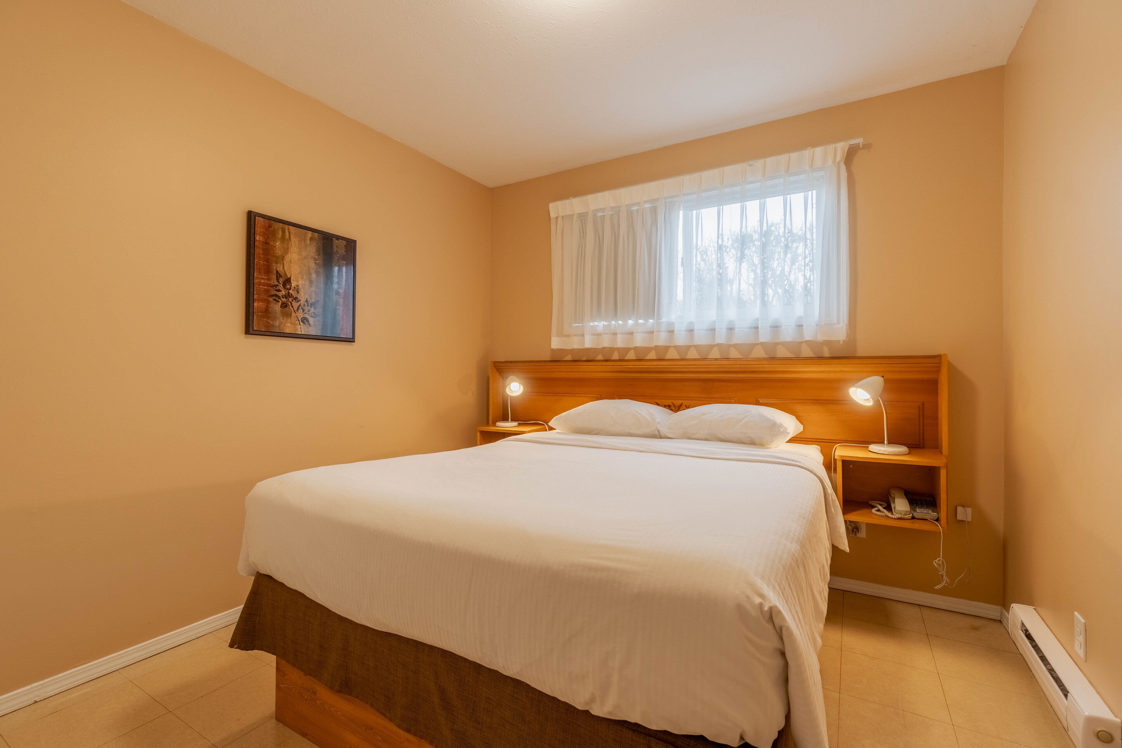 A bedroom with a bed, bedside lamps, a nightstand, a window with curtains, and a wall-mounted painting.