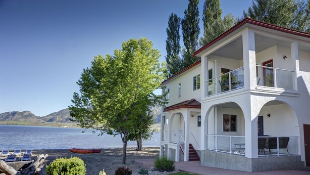 A two-story house with a balcony overlooking a lake, a red boat on the shore, and a well-maintained lawn. - Sandy Beach Suites (Osoyoos)