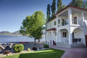Ein zweistöckiges Haus mit einem Balkon mit Blick auf einen See, ein rotes Boot am Ufer und eine gepflegte Rasenfläche.