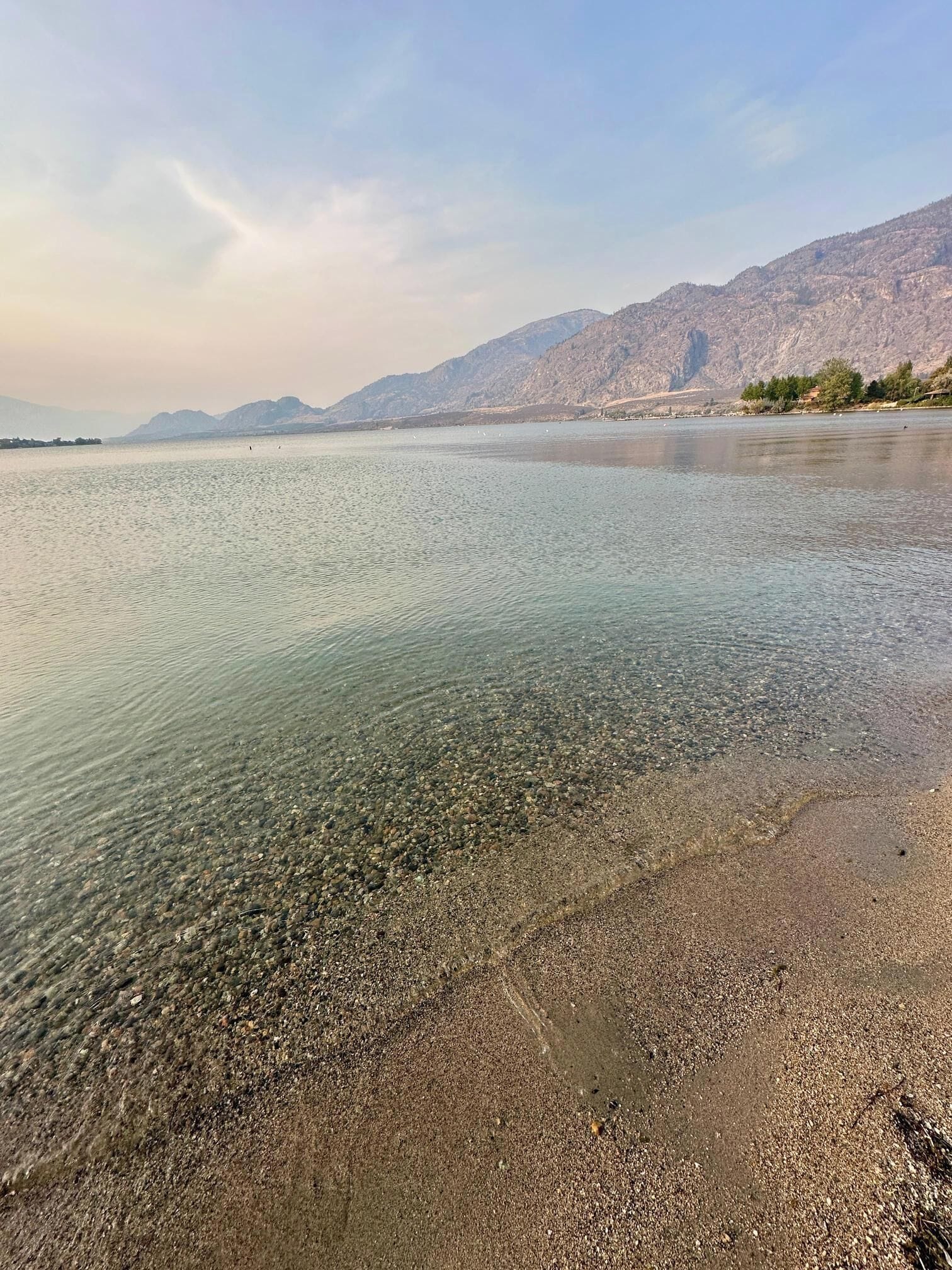 Un lac aux eaux limpides, bordé d’une plage de galets, avec des montagnes en toile de fond.
