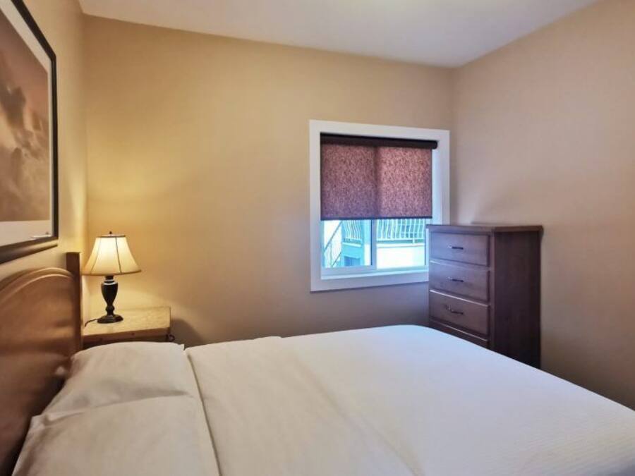 A hotel room with a bed, a nightstand with a lamp, a dresser, a window with blinds, and a framed picture on the wall.