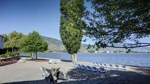 A beach with sun loungers and a tree, a building in the background, and a body of water. - Sandy Beach Suites (Osoyoos)