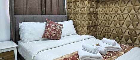 Cama bem arrumada com lençóis brancos e um edredom estampado em vermelho e dourado. Há duas toalhas enroladas sobre a cama. O quarto possui uma cabeceira com padrão geométrico e um criado-mudo branco com gaveta.