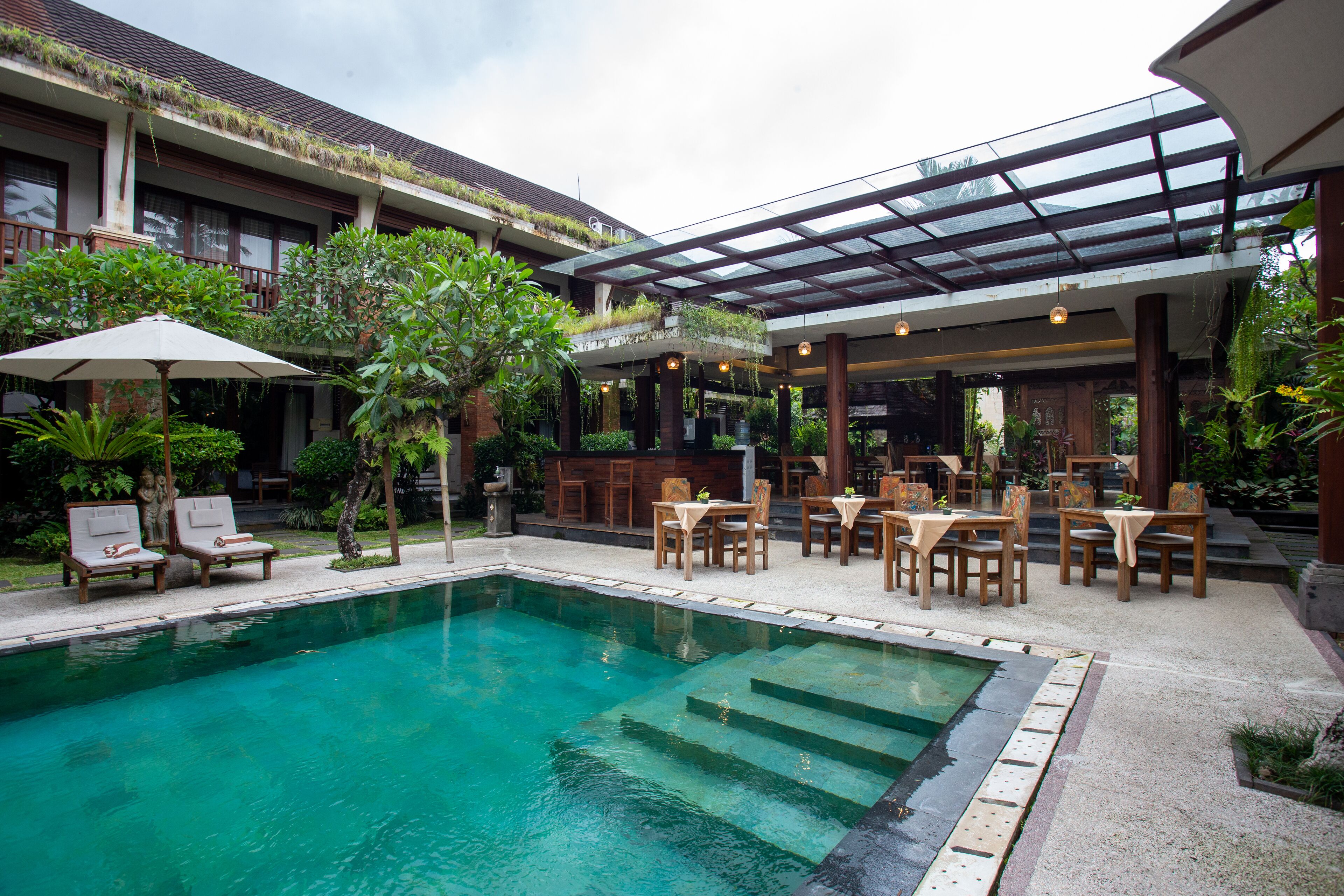 A swimming pool with lounge chairs and tables, surrounded by greenery and a covered patio area.