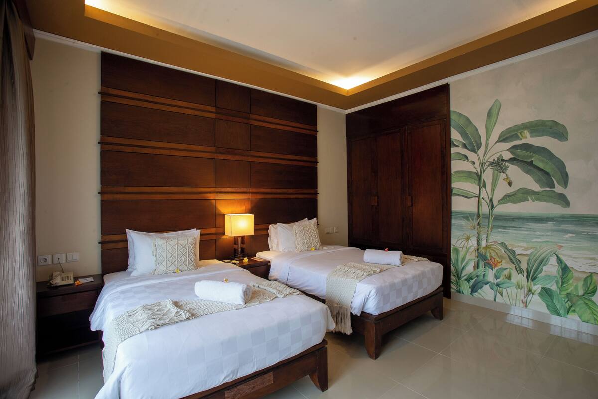 A hotel room with two beds, a nightstand, a lamp, a large wooden headboard, and a mural of a tropical scene.
