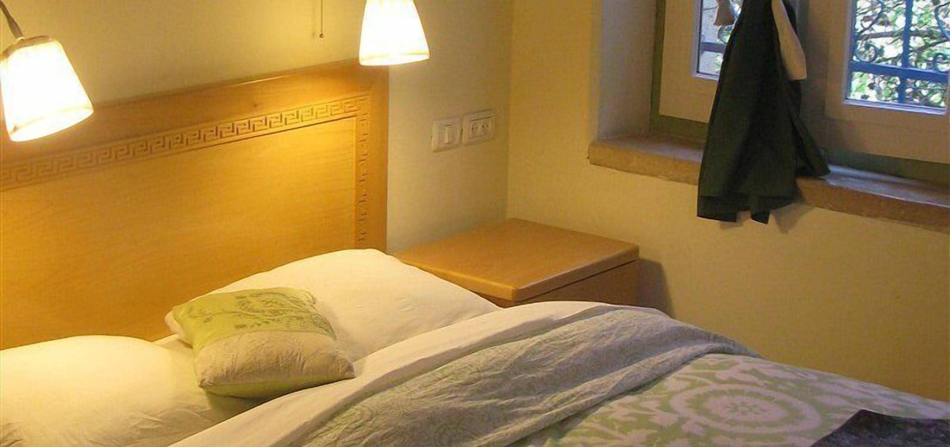 Un dormitorio con cama, almohadas, una mesita de noche y una ventana con cortina.