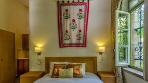 Quarto de hotel com uma cama, duas mesas de cabeceira, um criado-mudo, uma janela com cortinas e um tapeçário fixado na parede.