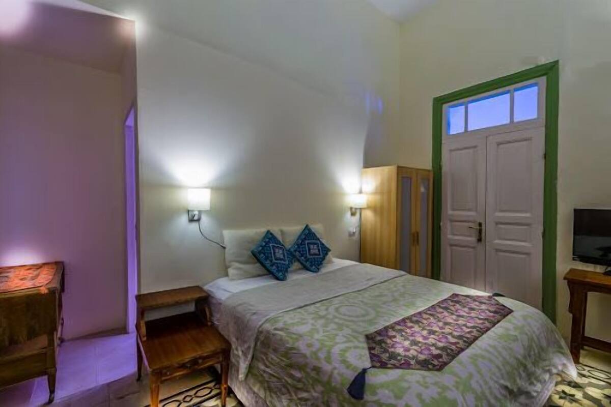 A bedroom with a bed, two wall-mounted lamps, a wooden nightstand, a television on a stand, and a door with a window.