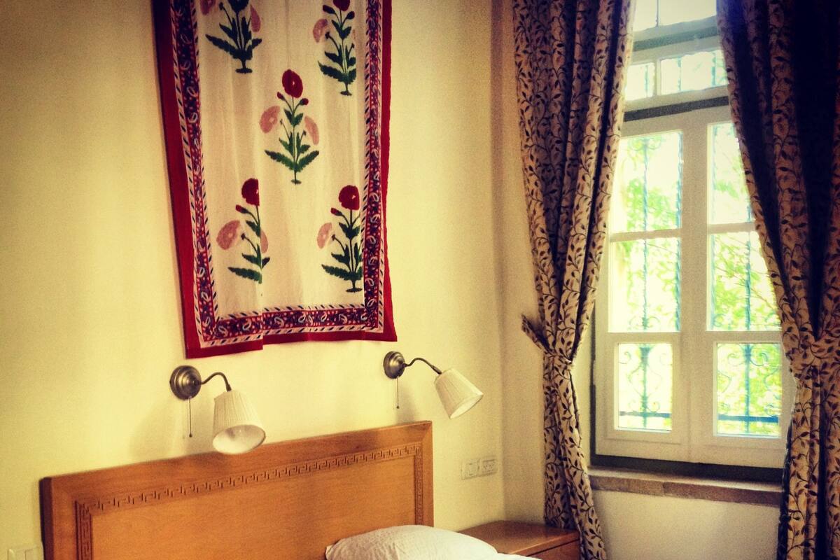 A bedroom with a wooden headboard, two bedside lamps, a bed with white linens, a patterned throw, and a window with curtains.