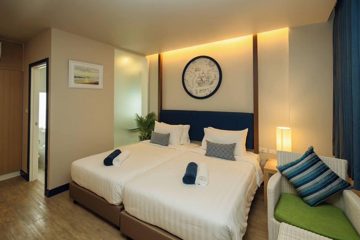 A hotel room with two beds, a large wall map, a side table with a lamp, and a green chair.