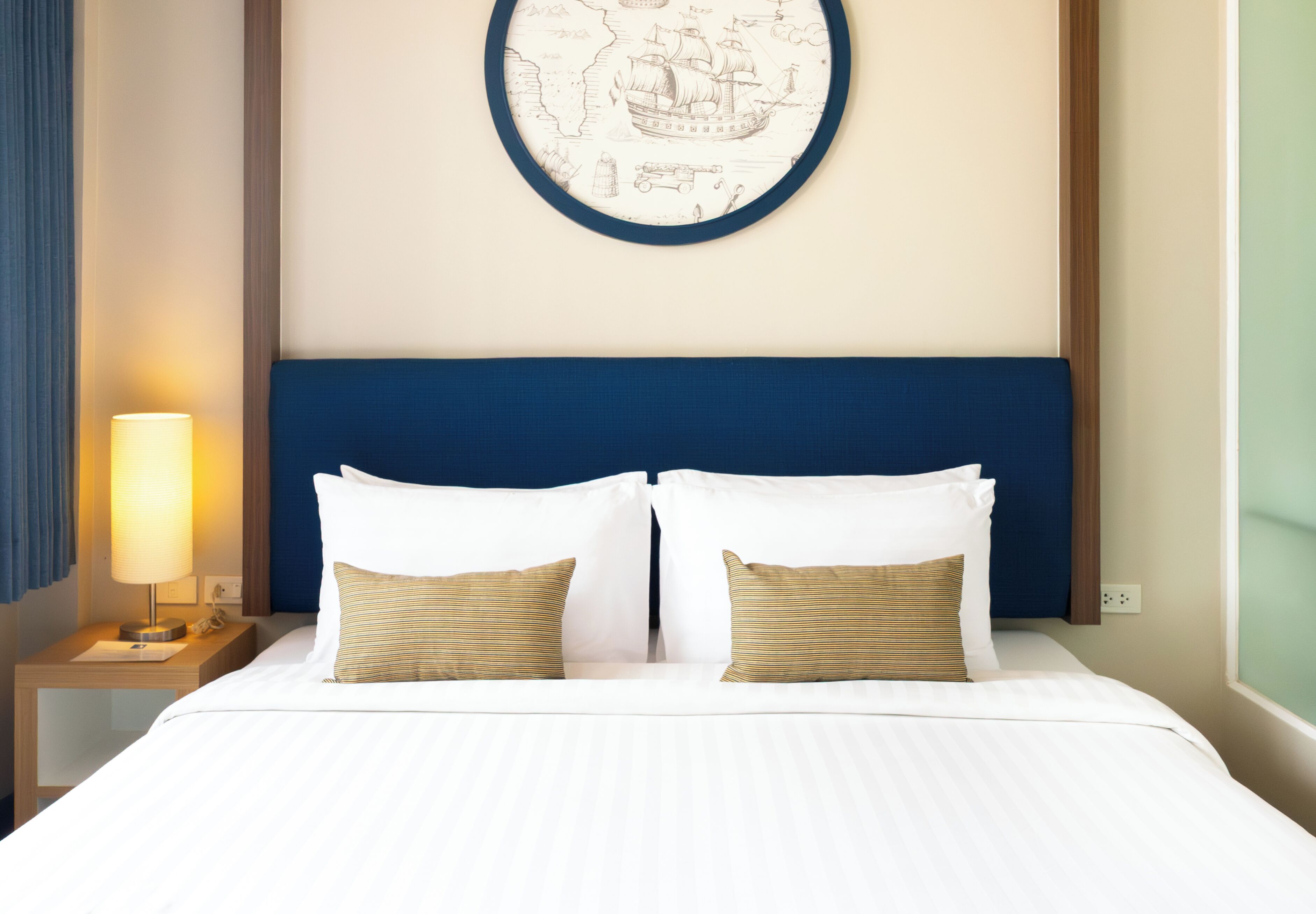 Una cama bien tendida con sábanas blancas y dos almohadas, una lámpara de noche y una obra de arte colgada en la pared que representa un barco de vela.
