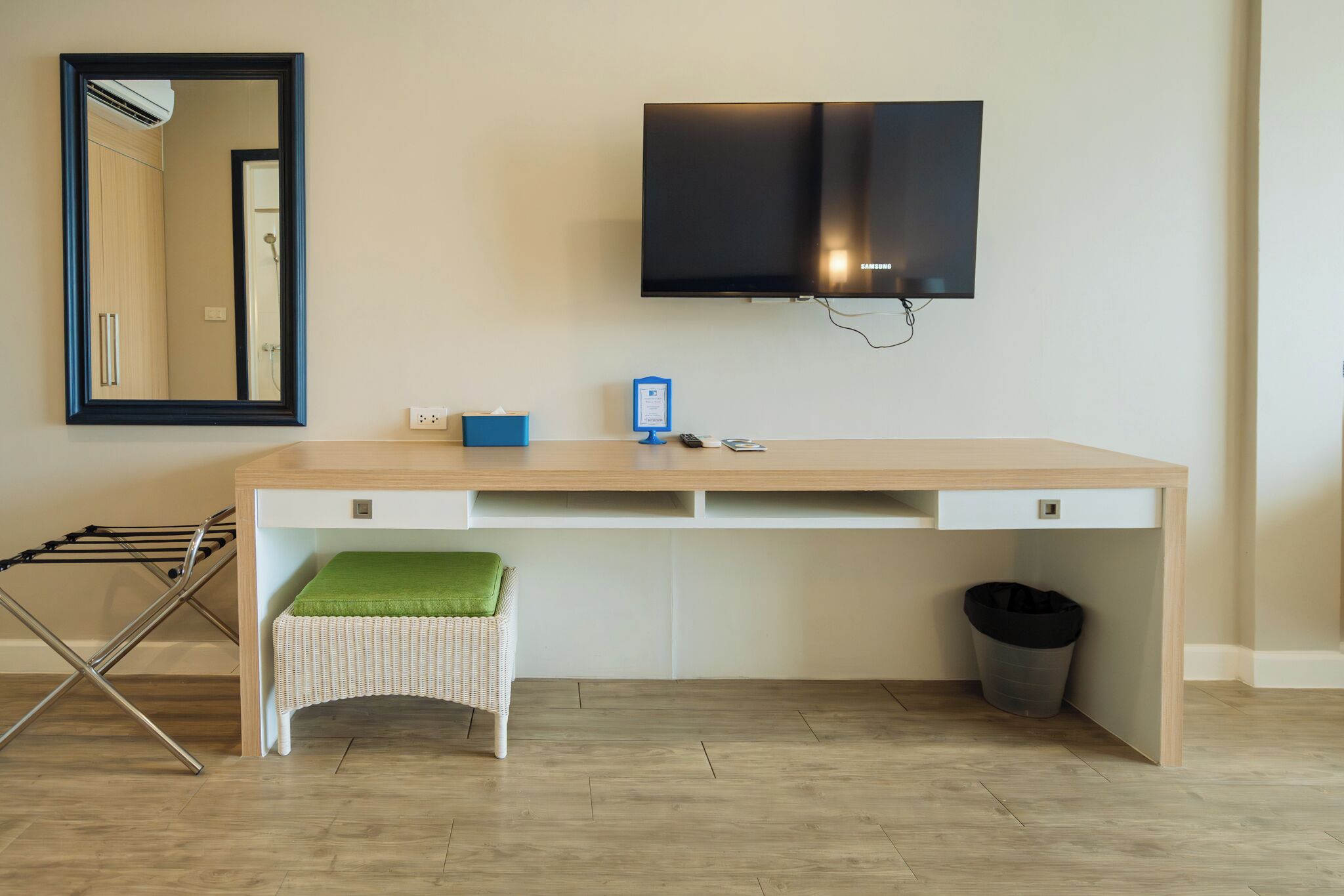 Una habitación moderna con un escritorio grande, un televisor montado en la pared, un espejo y una pequeña silla de mimbre.