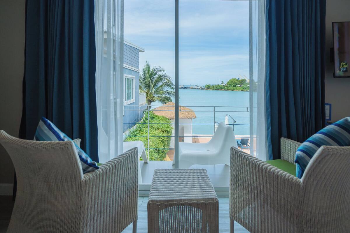 A balcony with wicker furniture, a white table, and a view of the sea and greenery.