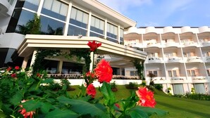 Un hotel de varios pisos con un vestíbulo cubierto, grandes ventanales y un césped bien cuidado con flores rojas.