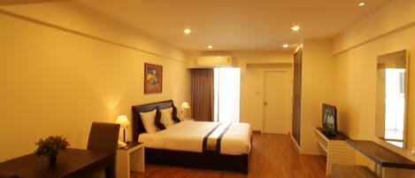 A hotel room with a large bed, a desk, a chair, a small table with a plant, and a TV on a stand.