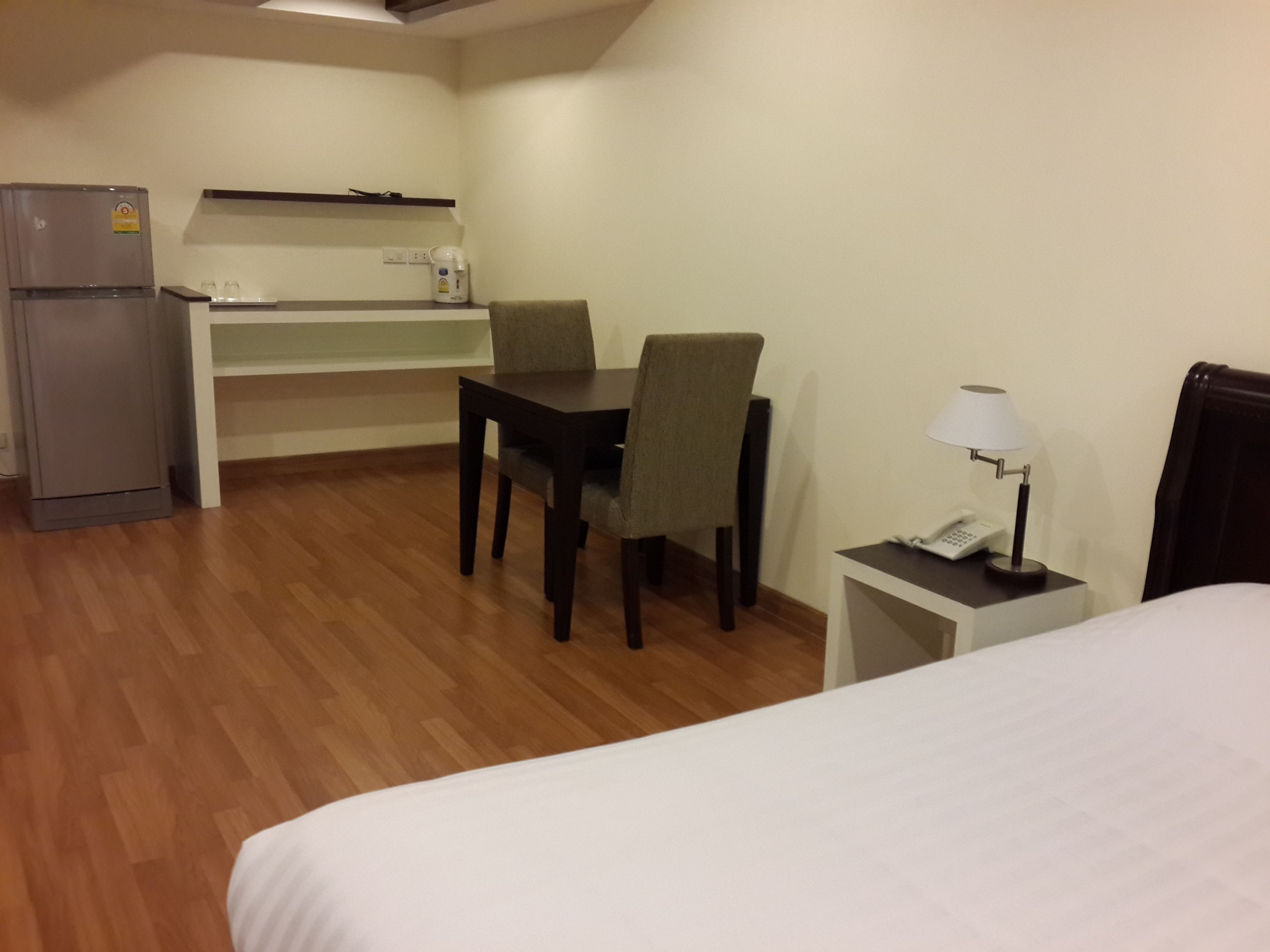 A hotel room with a bed, a small desk with a chair, a refrigerator, a coffee maker, and a bedside table with a lamp.