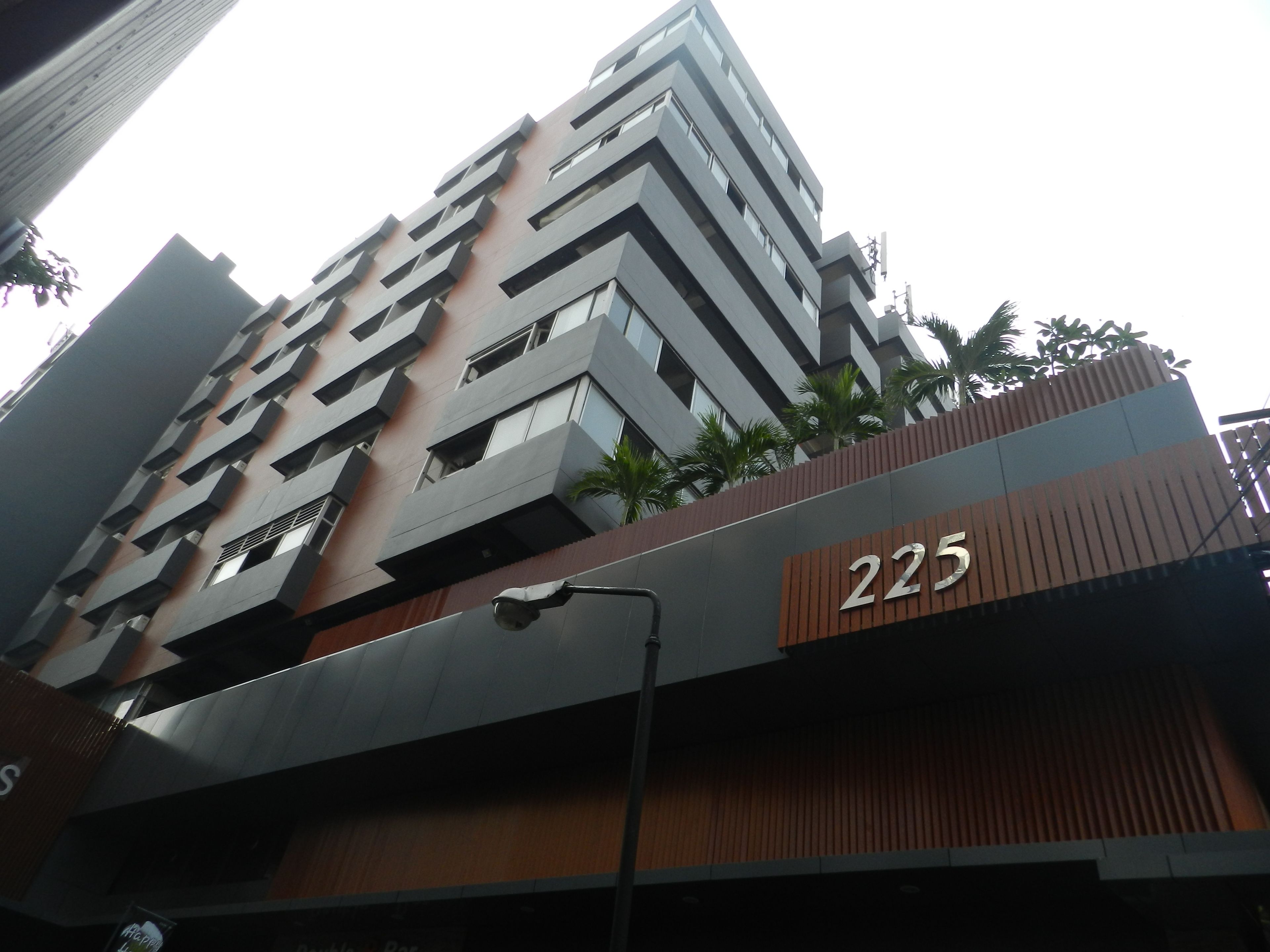 Un edificio moderno con el número '225' destacado en la fachada, que combina paneles de vidrio y madera, y un pequeño espacio verde con plantas.